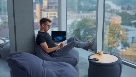 coming closer to a young man sitting relaxed near the window. man wearing glasses writing something in paper book. blurred backdrop.