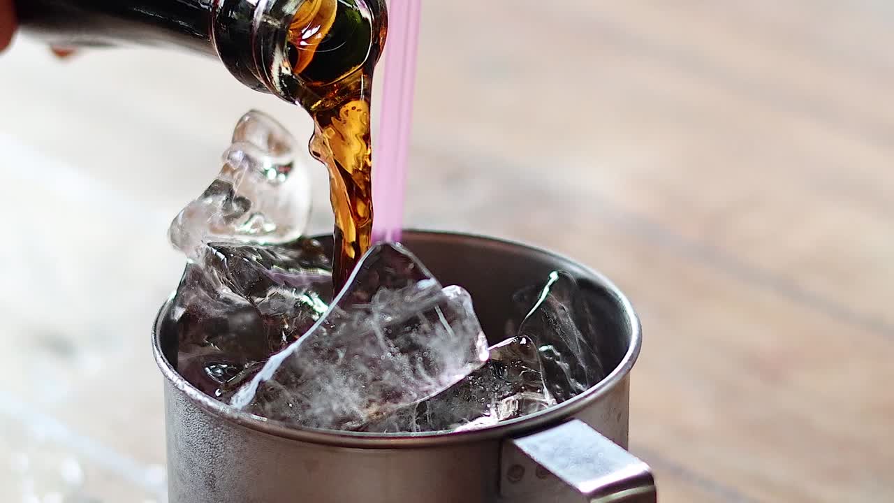 Close-up of soda being poured from a bottle into a metal cup filled with ice cubes.