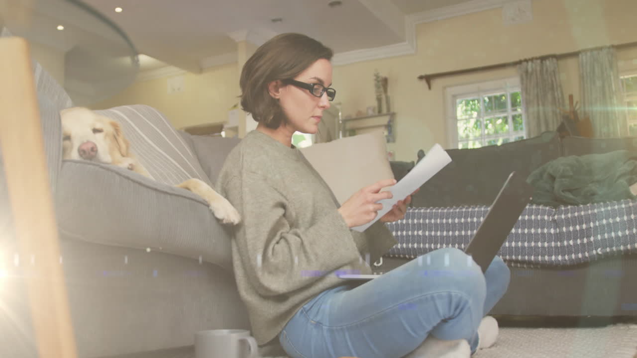 punto de luz contra mujer caucásica con computadora portátil leyendo un documento mientras el perro duerme en el sofá