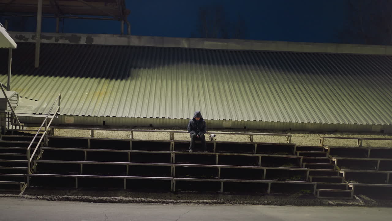 A man in a black outfit and jacket sits alone in an empty stadium, with a soccer ball beside him, with background dimly lit