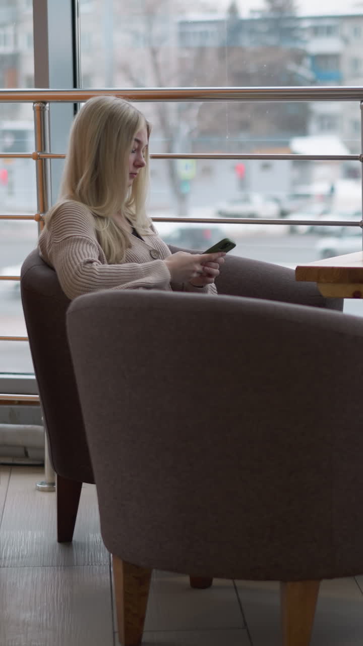 dama sentada sola en un acogedor espacio del centro comercial, absorta en su teléfono, con una vista de la ciudad ocupada a través de grandes ventanas, el momento tranquilo contrasta con la bulliciosa ciudad afuera