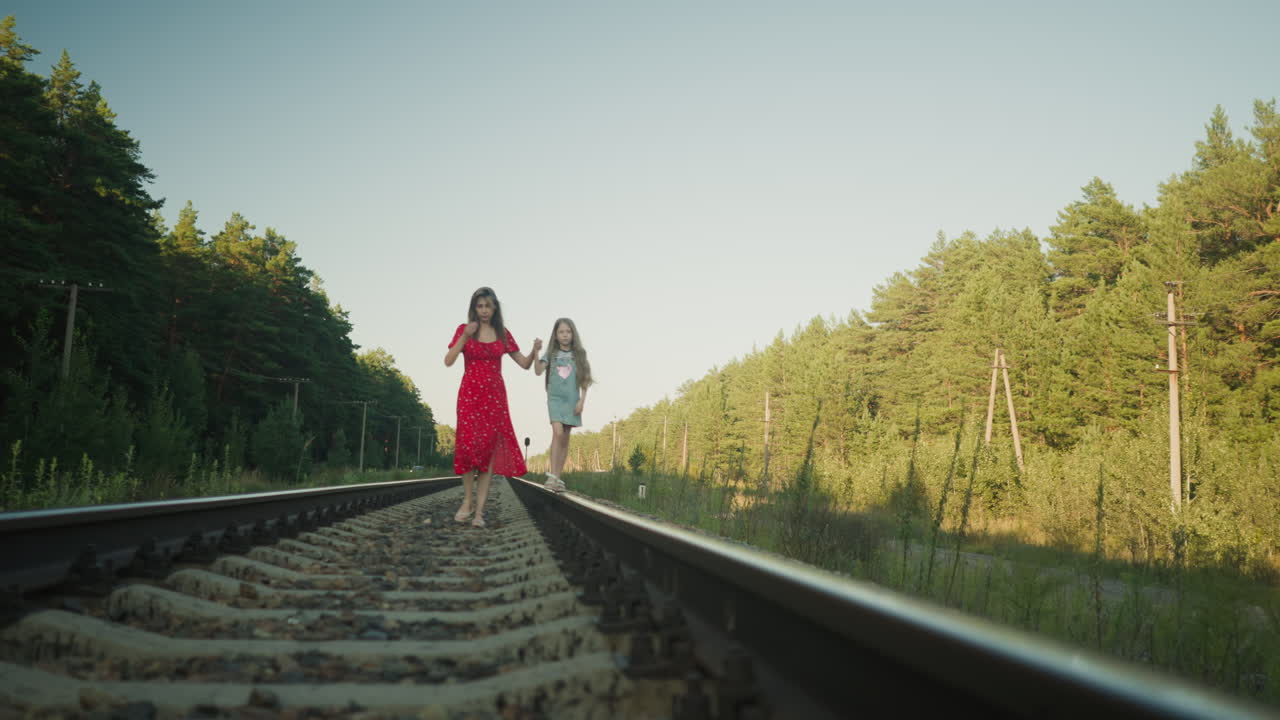 side view of mother in red dress walking closely with daughter who parks hair to back while balancing on rail beam along quiet track bordered by forest and power poles during sunny outdoor walk