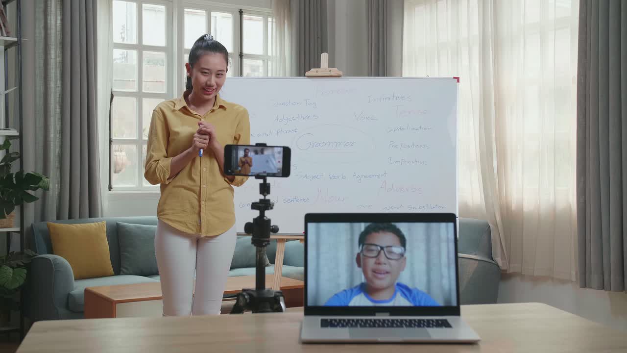 Asian Female Teacher Shoots Video By Smartphone Camera And Having Video Call On Laptop While Teaching English At Home