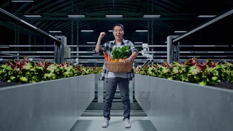 cuerpo lleno de feliz agricultor masculino asiático con canasta de verduras celebrando mientras estaba de pie en la granja inteligente