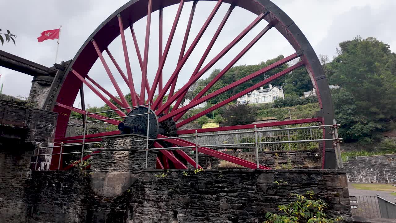 A large red water wheel dominates a stone platform atop a hillside, with surrounding railings
