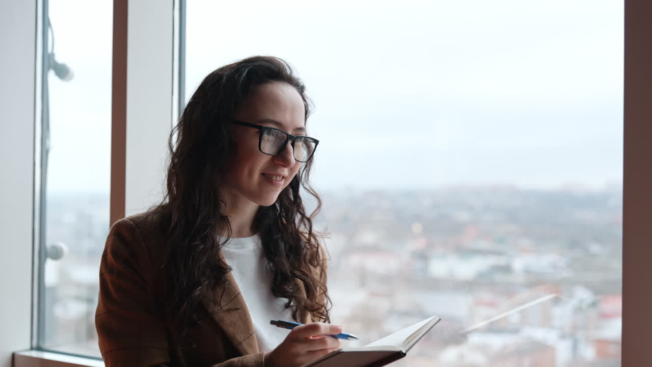 Long-haired brunette in glasses writes down into her paper book. Colleague pregnant lady drinks coffee and speaks to her co-worker.