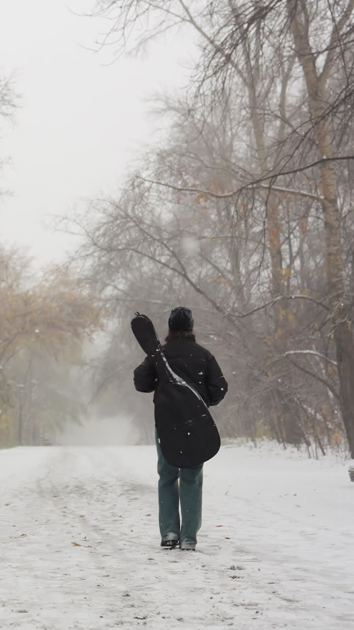 기타 을 들고 눈 인 공원에서 걸어가는 소녀의 뒷면, 길을 가로지르는 겨울의 나무와 벤치, 조용한 눈이 내린 날을 포착합니다.