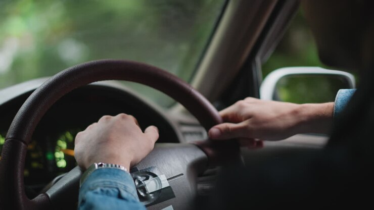mani sul volante all'interno dell'auto, guidatore che percorre una strada alberata con manica di jeans e orologio argentato visibili, luci del cruscotto accese, bokeh delicato attraverso il finestrino bagnato, tragitto casa-lavoro cinematografico e suggestivo