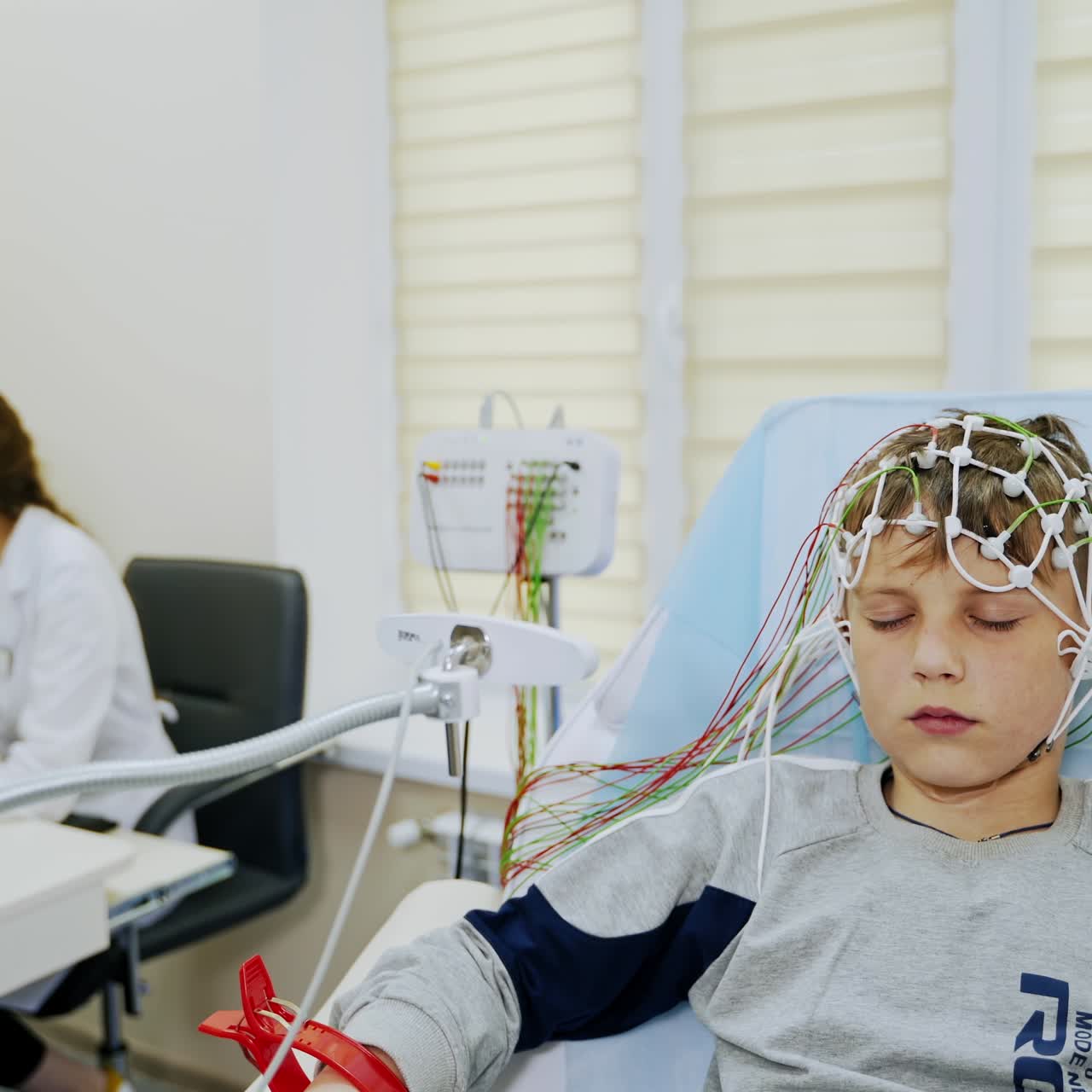 Young boy sits in a chair with lots of sensors attached to his head. Female doctor sits at computer screen at backdrop