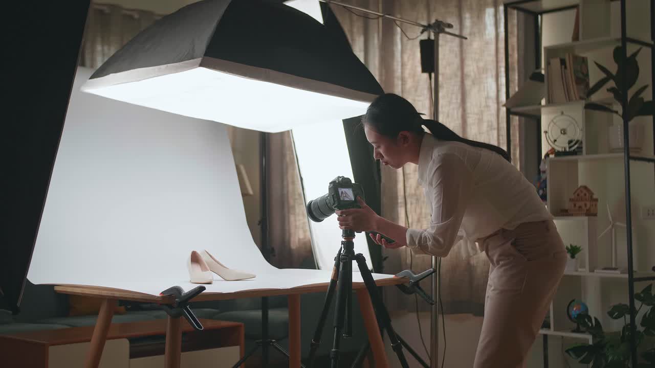 fotógrafa asiática hablando en su teléfono inteligente mientras toma fotos de zapatos femeninos en un estudio casero