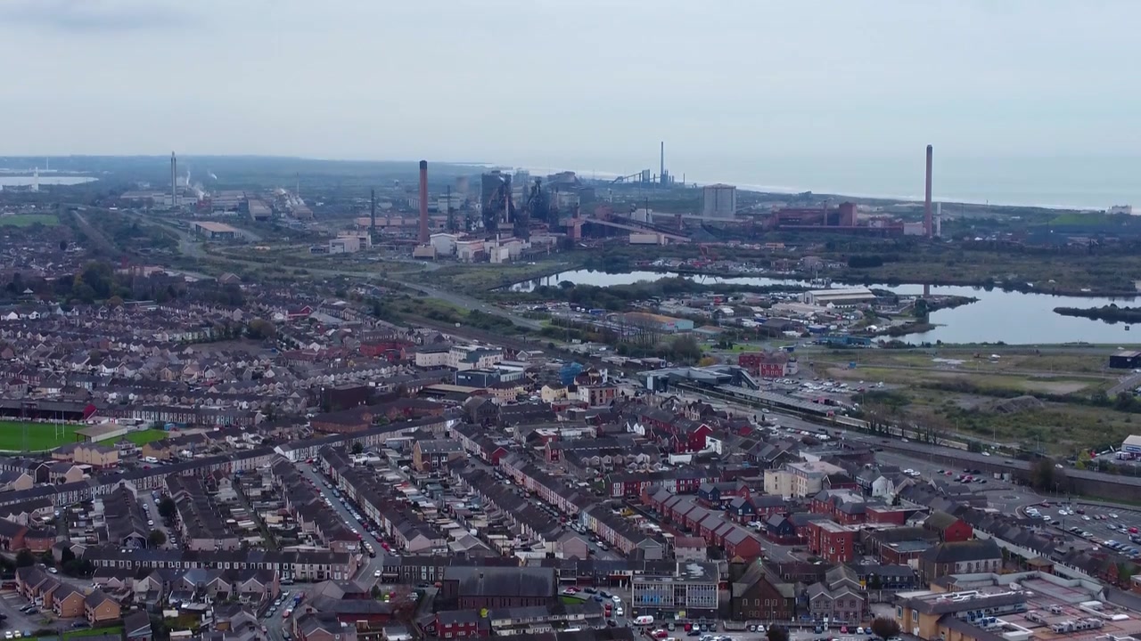 Hurtig luftoptagelse med tilt op over travl motorvej med trafik og huse foran stålværk i Port Talbot med mørk overskyet himmel 4K