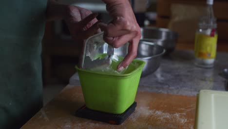 Glass-container-of-plain-white-flour-tipped-into-green-plastic-container-and-measured-on-scale,-filmed-as-medium-closeup-slow-motion-shot