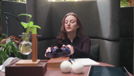 lady with brown hair removes headset and closes book while leaning back on cushioned seat, appearing relaxed and reflective in modern workspace with indoor plant and warm lighting details