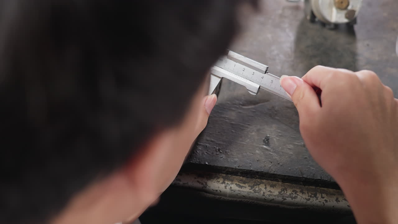 Vista dall'alto di un tecnico di laboratorio che usa un calibro a corsoio per misurare il diametro di un boris su un banco da lavoro sotto una luce fioca, mentre registra le misurazioni su un tablet vicino. Ambiente industriale con strumento di precisione.