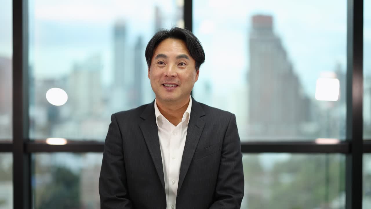 Asian businessman in suit smiles and waves, natural light, cityscape background, steady camera, professional mood
