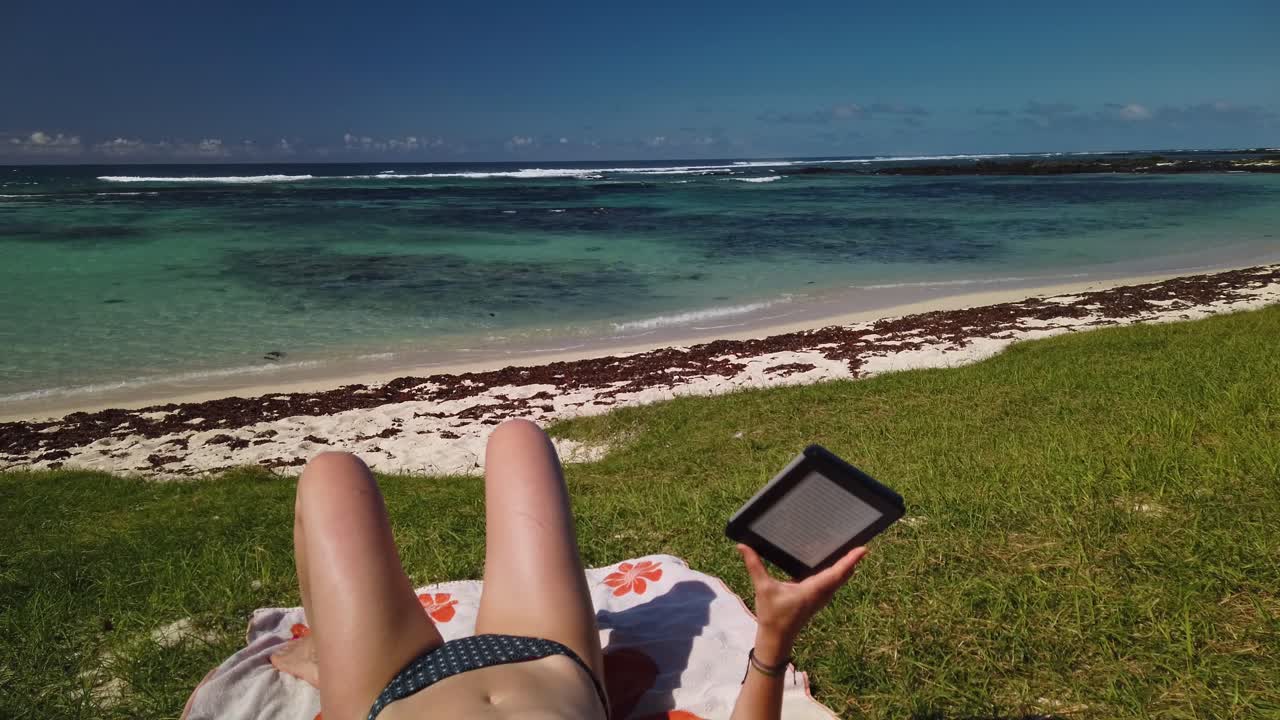 una toma que muestra las piernas, el abdomen y los brazos de una mujer que sostiene un libro electrónico en una playa tropical