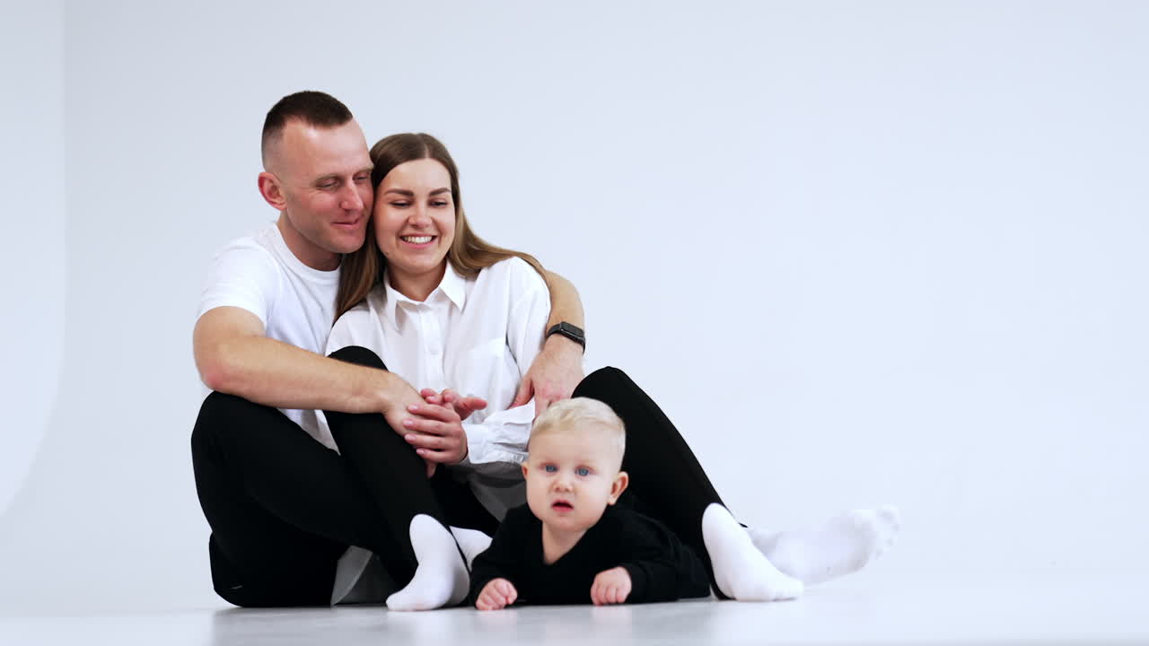 Caucasian couple sits on the studio floor embracing and looking at their baby. Calm child lies near the parents looking ahead attentively. White backdrop.
