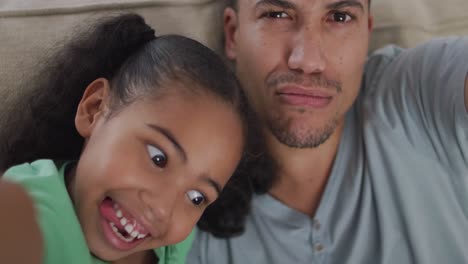 Happy-biracial-father-and-daughter-sitting-on-sofa-taking-selfies