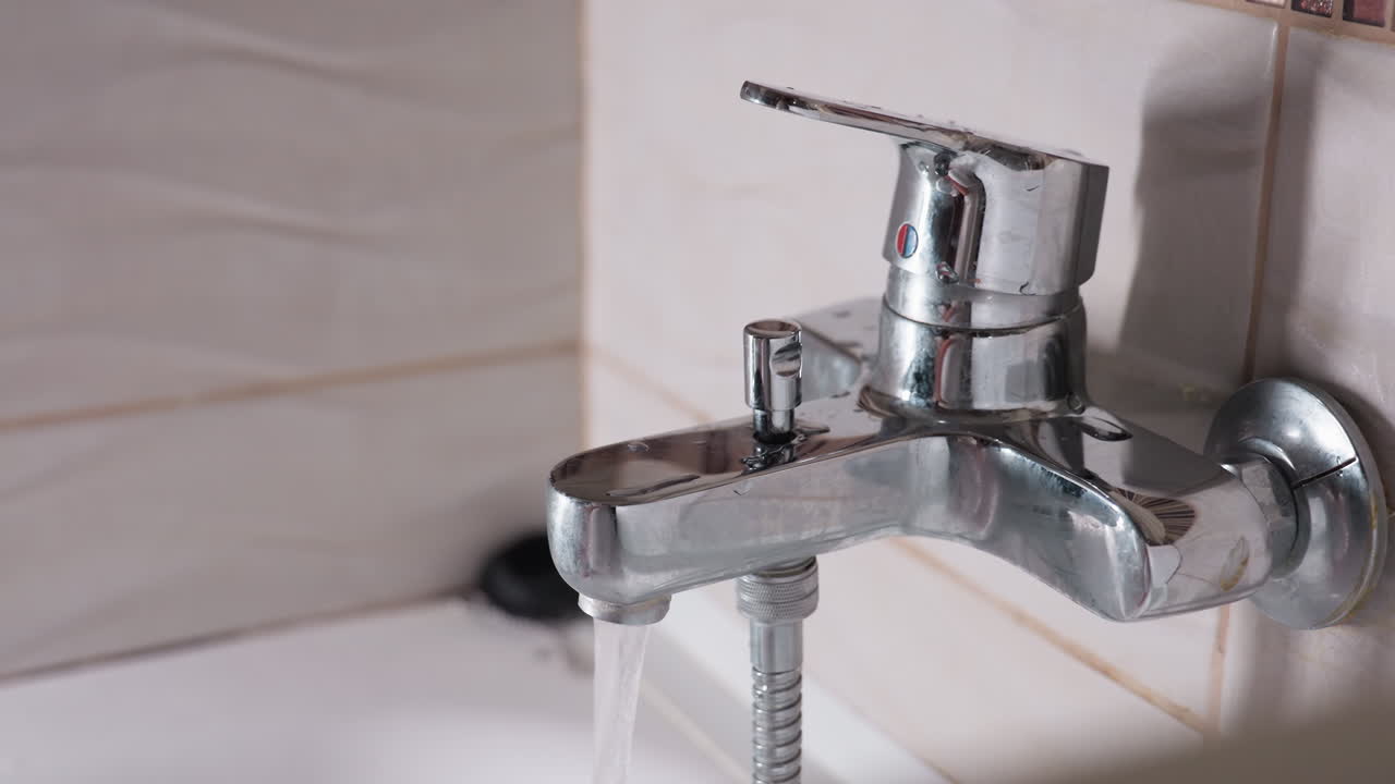 Hand view lady turning on shower tap, on chrome bath mixer, fingers twist knob, hose visible, soft light across tiles, bathroom setting, routine hygiene start, calm motion before washing