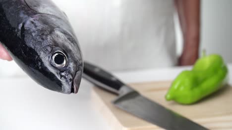 Chef-Sosteniendo-Un-Pescado-Fresco,-Cuchillo-Y-Pimienta-En-Una-Tabla-De-Corte-En-El-Fondo,-Delantal-Blanco