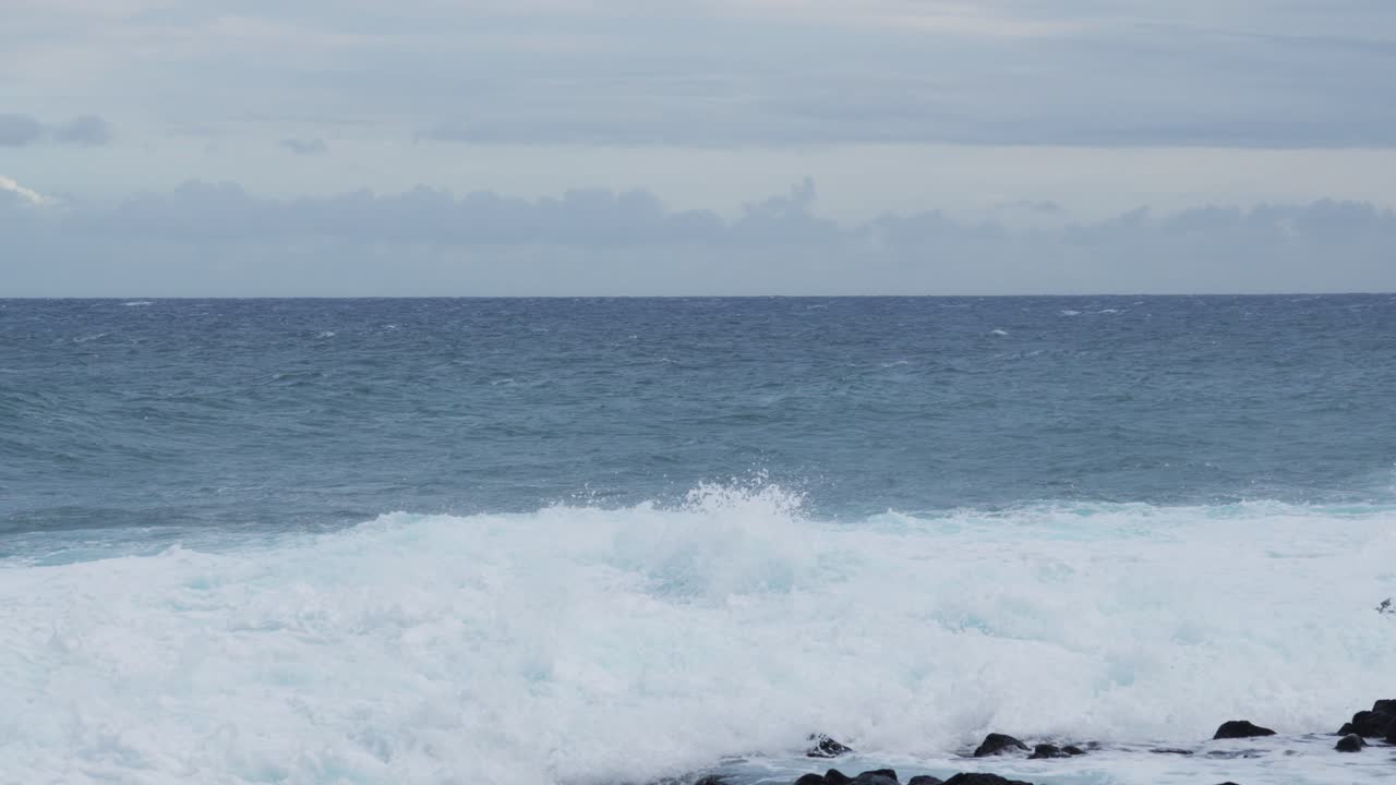 Wide ocean view with rolling waves crashing against black coastal rocks, foamy white surf spreading across the rugged shoreline beneath a cloudy sky