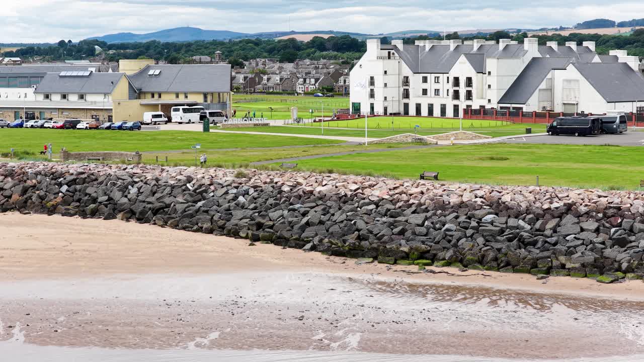 Drone glides over rocky shoreline toward golf clubhouse, green fairway, and town under daylight