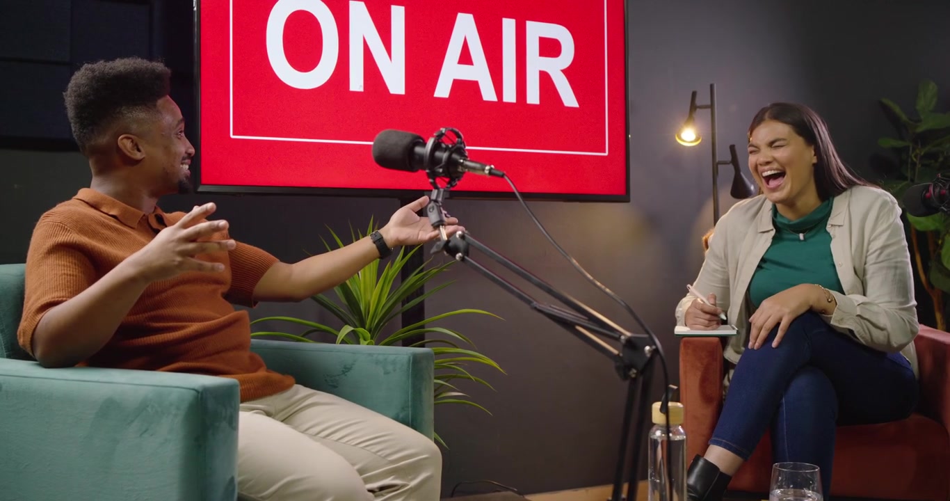Afrikansk kvinnlig programledare intervjuar manlig gäst, "ON AIR"-skylt lyser, mikrofoner, programledare skriver.