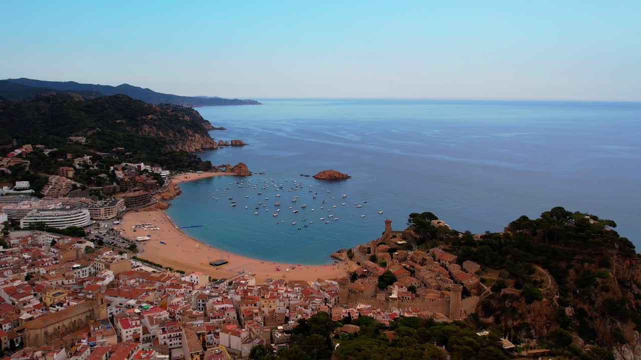 An aerial view of Tossa Del Mar in Spain dollying forward over the hills into the city, revealing the bay, sailboats, and oceanfront in a breathtaking coastal panorama.