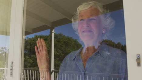 Happy-caucasian-senior-woman-at-home,-looking-out-of-window-on-sunny-day-smiling-and-waving