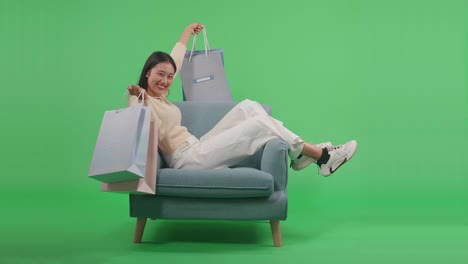 an asian shopping woman holding shopping bags up smile to camera while sitting on sofa in front of green screen