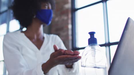 african american woman wearing face mask sitting in cafe with laptop disinfecting hands