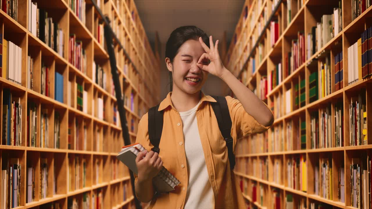 estudiante asiática con una mochila y algunos libros sonriendo y mostrando el signo de la mano de acuerdo sobre su ojo mientras estaba de pie en la biblioteca