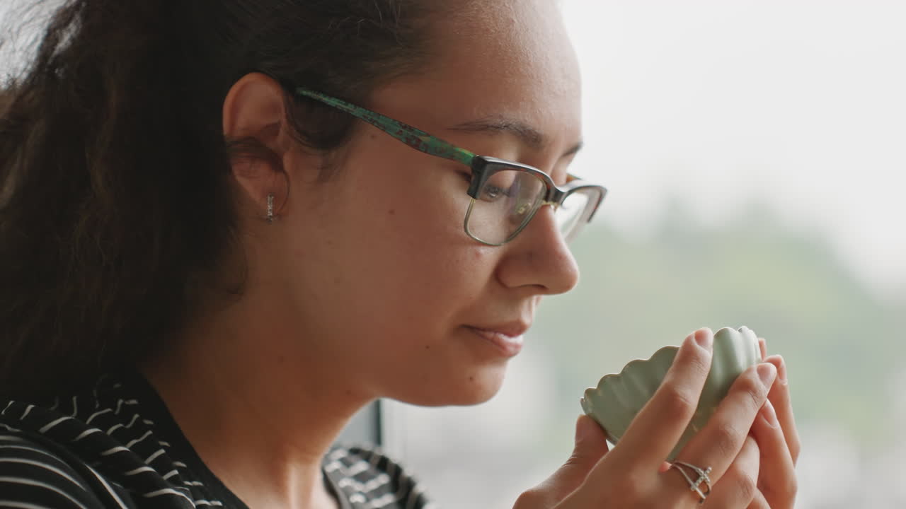 Donna che si gode la vista dalla finestra dell'hotel con il tè, inquadratura di profilo contemplativa, paesaggio urbano sfocato all'esterno, sereno inspirare dell'aroma, una pausa tranquilla per un lavoratore a distanza o uno scrittore, atmosfera di luce diurna morbida.