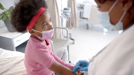 African-american-female-doctor-with-face-mask-making-injection-to-girl-in-hospital-room,-slow-motion