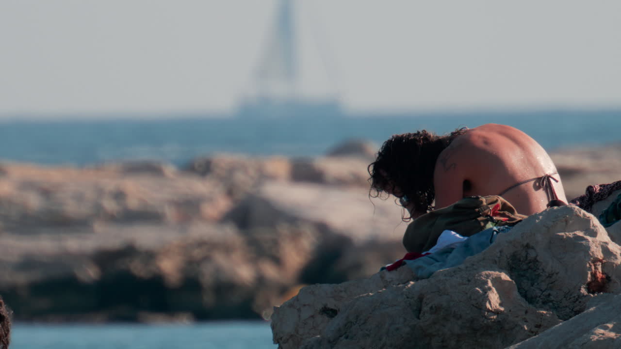 Cannes, France - October 10, 2025: A young woman with wet hair relaxes on seaside rocks under the warm sun, with the sea and a yacht in the distance