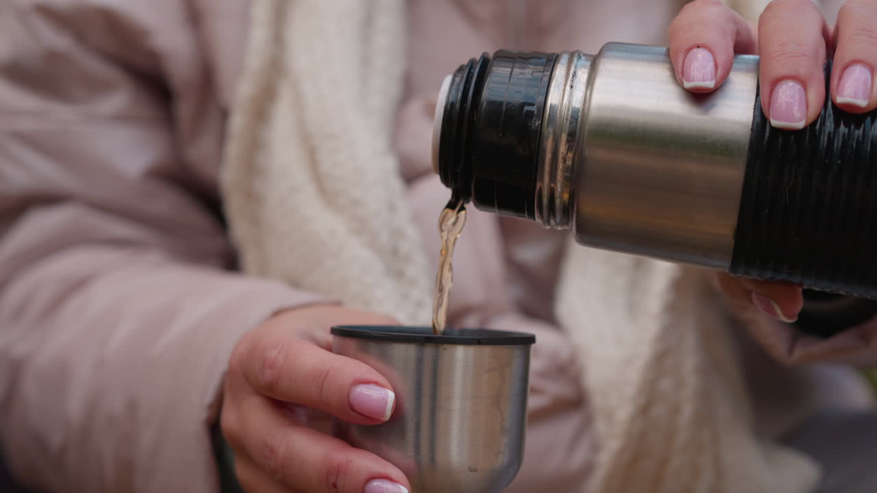 Detailed close up of feminine hands with fresh pink French manicure slowly pouring steaming herbal tea from stainless steel flask into small metal cup during outdoor autumn picnic