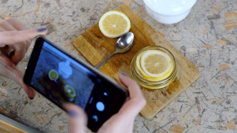 close-up of woman hand with phone making photo slices of lemon in jar.