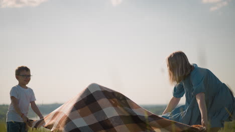 a woman in a blue dress and her son hold a large plaid scarf, playfully covering the younger son as they stand on a breezy hilltop. the scenic landscape and lake in the background