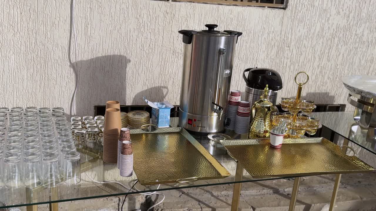 Servizio da caffè e tè arabo con macchina per l'acqua calda, caffettiera dallah, tazze da tè in vetro, bicchieri usa e getta e vassoi dorati su un tavolo da buffet, postazione di accoglienza per eventi interni