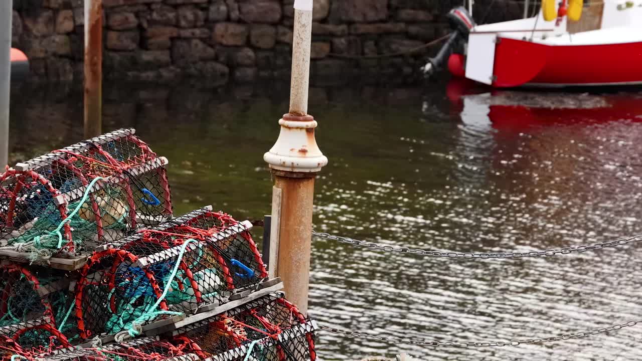A red boat is docked near a stone wall with stacked fishing traps in the foreground.