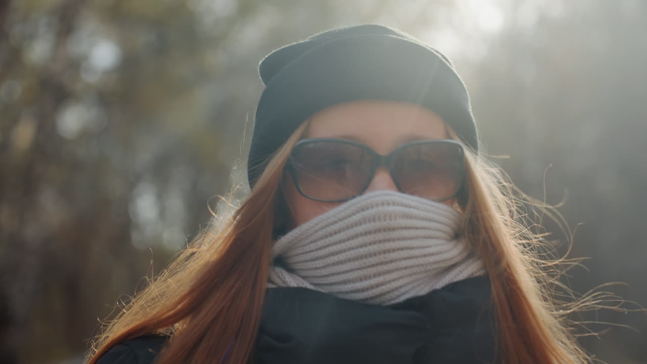 Masked white woman in forest backlit by autumn sun, beanie and scarf hiding lower face, sunglasses and backpack, slow cautious walk suggesting prowler or snatcher, cinematic moody tension with closeup