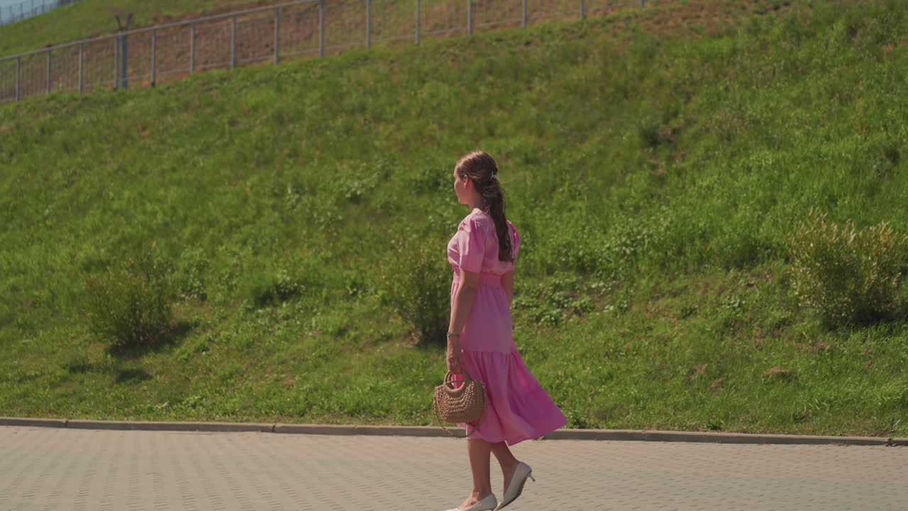 Donna che cammina in abito rosa lungo un terrapieno erboso, la luce del sole proietta ombre soffici, che trasporta un piccolo bouquet, tacchi casual, lunga treccia, sereno pomeriggio estivo, sentiero del parco urbano, movimento naturale