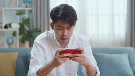 Asian Man Playing Game On Smartphone And Smiling While Sitting On Sofa In The Living Room
