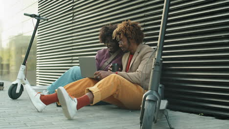 african american friends sitting on street, using laptop and chatting