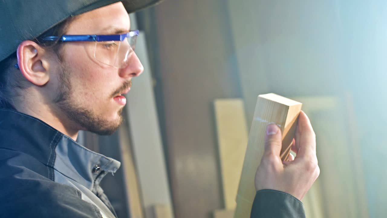 고글과 모자를 입은 집중된 젊은 수염이 있는 목공이 작업 조각을 확인합니다.