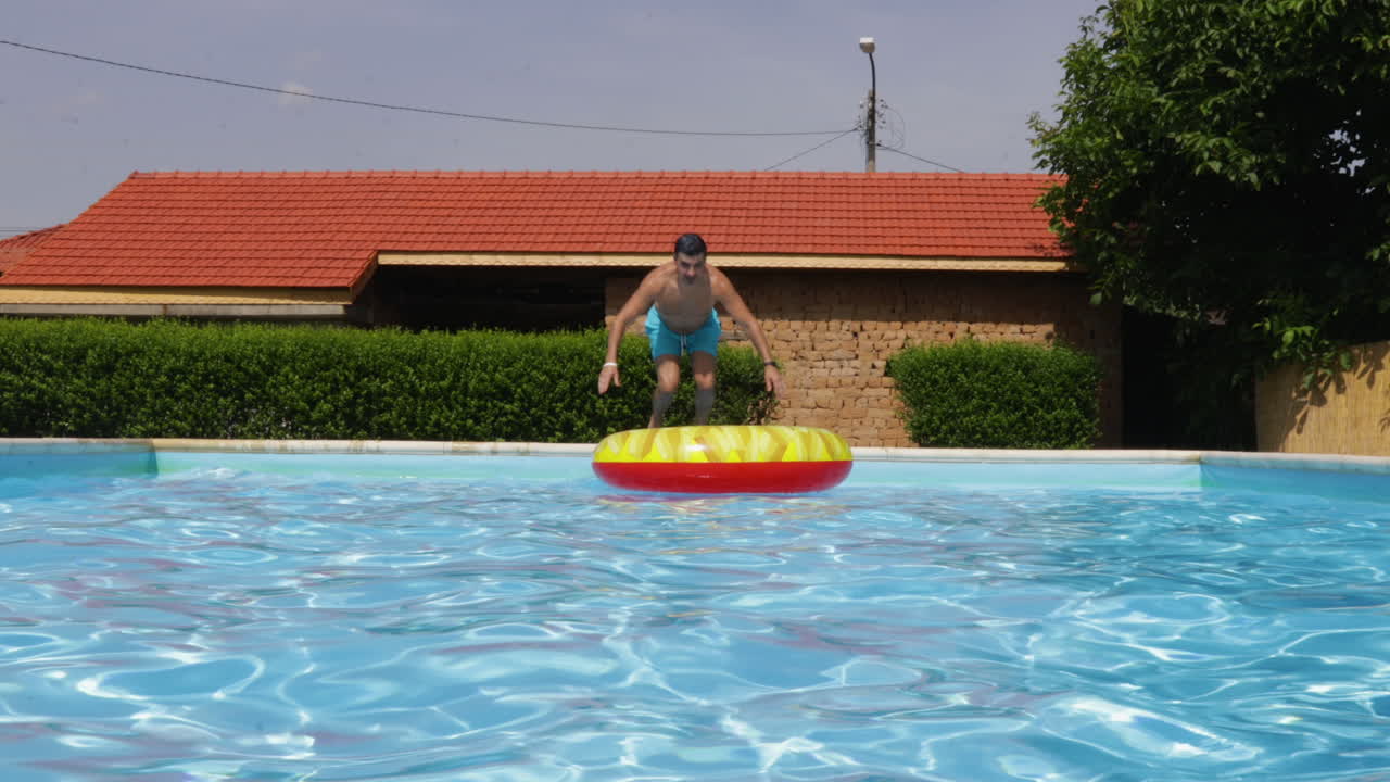 hombre llevando un inflatable rojo camina junto a una piscina en un día soleado, preparándose para saltar al agua