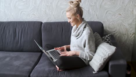 Cheerful-young-blond-woman-sitting-on-couch-in-living-room-and-using-laptop