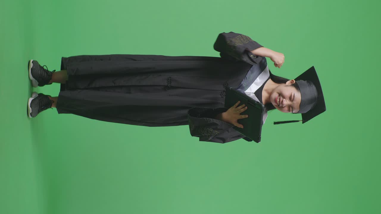 Studentessa asiatica laureata a figura intera, con tocco e toga, che guarda un tablet e poi esulta, su sfondo a schermo verde in studio