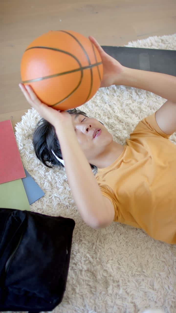 Video verticale: Ragazzo adolescente asiatico sdraiato sul pavimento che tiene in mano un pallone da basket, si rilassa a casa con dei libri vicini