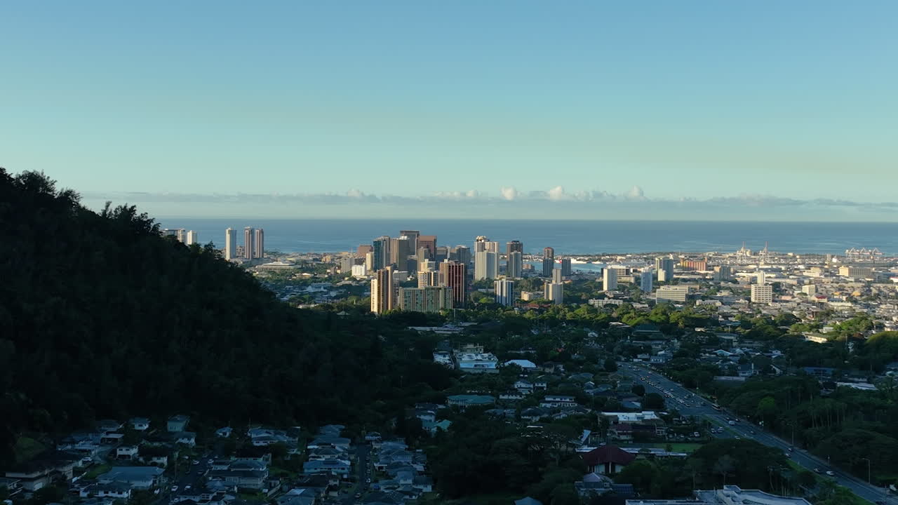 Downtown skyline of Honolulu City during golden sunrise. Suburb neighborhood in Hawaii. Aerial descend wide shot. Busy traffic on highway. Blue seascape in distance.